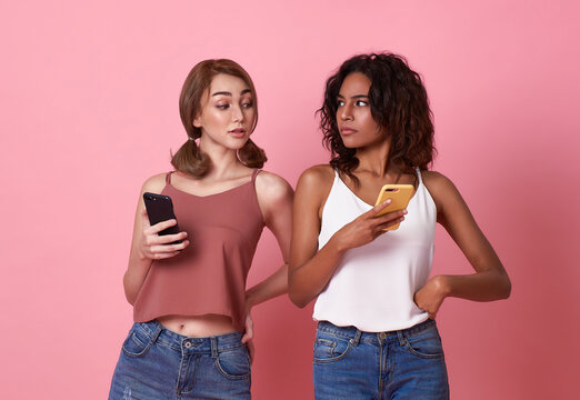 Young two women using mobile phone while brunette woman looks at smartphone her friend on pink background. secret privacy in social media.