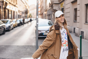 Happy girl with blonde hair in light cap and brown trench coat with handbag smiling outdoors, turn around and smiling. Stylish girl in fashion outfit walking over the city, look happy, hold bag.