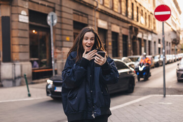 Happy smiling brunette woman looking at smartphone in her hand with excited face expression, unable...