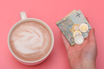 Kawa z pianką w filiżance obok dłoni z pieniędzmi na różowym tle