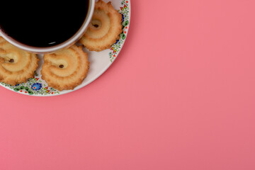 Filiżanka z mocną kawą po lewej stronie zdjęcia na różowym tle, na spodku leżą ciasteczka