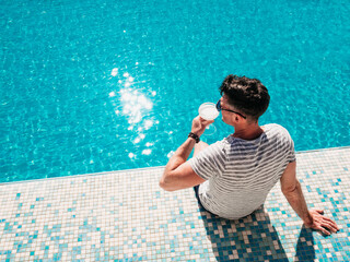 Handsome man with a glass of drink sits near the swimming pool of a cruise ship. Sunny morning,...