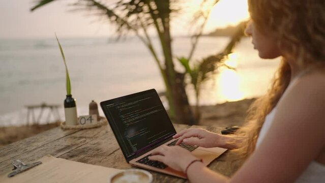 Young female developer working on laptop by the ocean. Woman freelancer coding at outdoor tropical cafe at sunset. Caucasian girl working remotely typing on computer at exotic location. Worldwide work