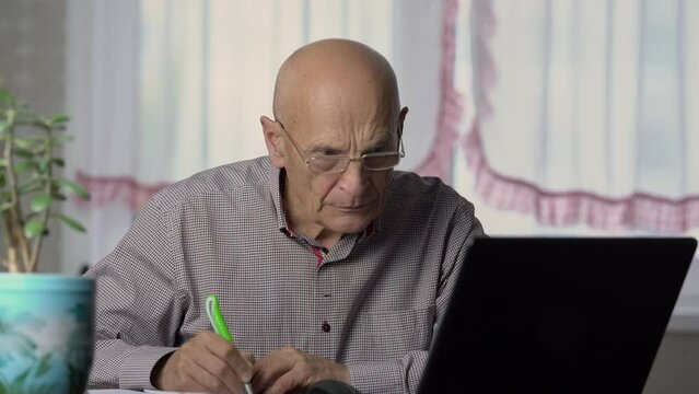 Senior man writes necessary information in paper notebook from laptop. Elderly man focuses on finding interesting information in internet
