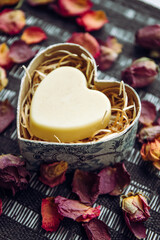Solid Body cream bar in a heart shape gift box on black background, decorated with dry rose flower petals indoors.