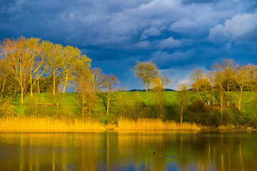 Sunlit spring landscape at lake and dark blue sky clouds in background
