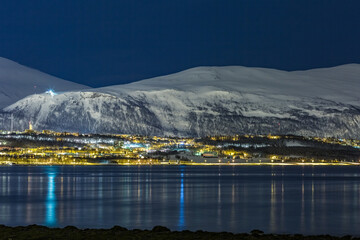 Long exposure cold night photograph of the Norwegian city of Troms&oslash;, Northern Norway. 30 seconds. Mountains covered in snow and motion blurred water reflections of the lights.