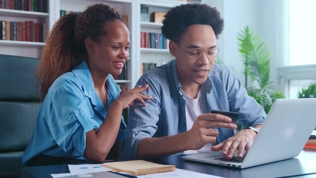 Young cheerful African American man and woman make large purchase together through laptop and use credit card to pay for goods in online store or renew subscription sits at table in home office
