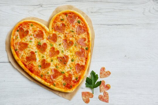 Heart shaped pizza with heart shaped salami on parchment paper with white wood background.Creative art food idea for Valentine or Mother day.Top view.Copy space