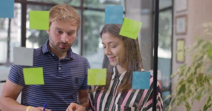 Two work colleagues organizing startup project ideas. Team of caucasian business people standing near glass window with sticky notes. Scrum methodology with colorful stickers.