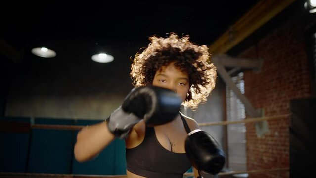 Afro american woman boxer in boxing gloves looks at camera stands on boxing ring and boxing. Sportswoman training in gym. Girl's power, woman train workout, sport fight, self-defense and feminism.