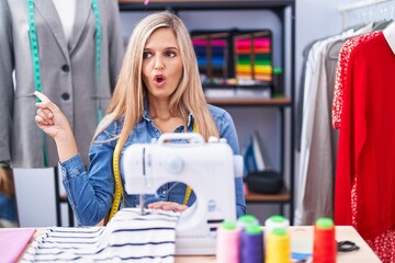 Blonde woman dressmaker designer using sew machine surprised pointing with finger to the side, open mouth amazed expression.