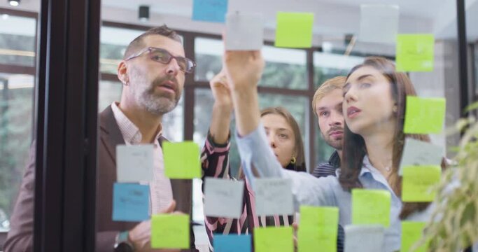 Work colleagues discussing startup project ideas. Team of caucasian business people standing at glass wall with sticky notes. Scrum methodology with colorful stickers. Left camera movement.