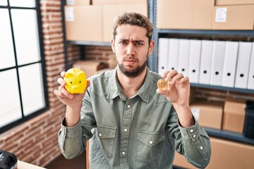Hispanic man with beard working at small business ecommerce holding piggy bank and bitcoin skeptic and nervous, frowning upset because of problem. negative person.