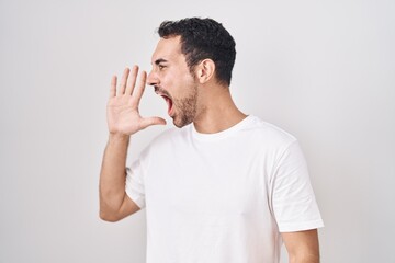 Handsome hispanic man standing over white background shouting and screaming loud to side with hand on mouth. communication concept.