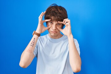 Hispanic young man standing over blue background trying to open eyes with fingers, sleepy and tired for morning fatigue