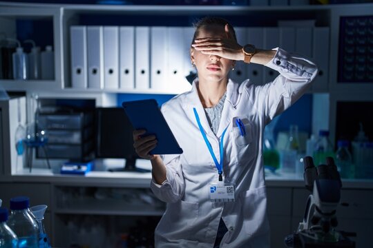 Beautiful blonde woman working at scientist laboratory late at night covering eyes with hand, looking serious and sad. sightless, hiding and rejection concept