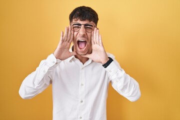 Young hispanic man standing over yellow background shouting angry out loud with hands over mouth