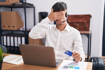 Young hispanic man working using computer laptop holding credit card covering eyes with hand,...