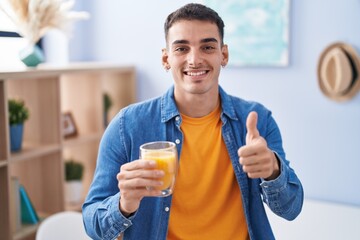 Handsome hispanic man drinking glass of orange juice smiling happy and positive, thumb up doing excellent and approval sign