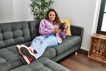 Obraz premium Young beautiful hispanic woman using touchpad lying on sofa at home