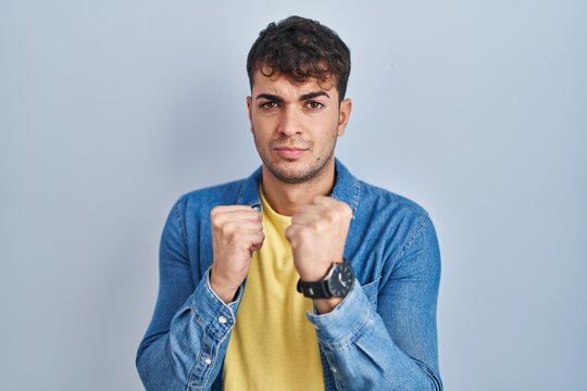 Young hispanic man standing over blue background ready to fight with fist defense gesture, angry and upset face, afraid of problem