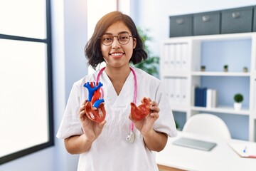 Young latin woman wearing doctor uniform holding anatomical model of heart at clinic