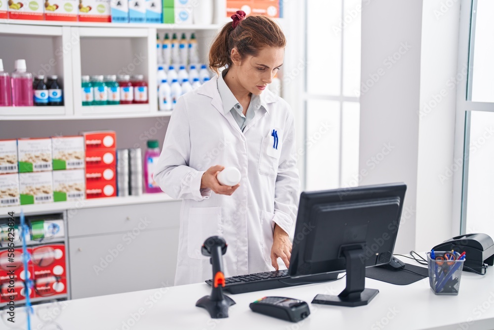 Poster Young woman pharmacist holding pills bottle using computer at pharmacy