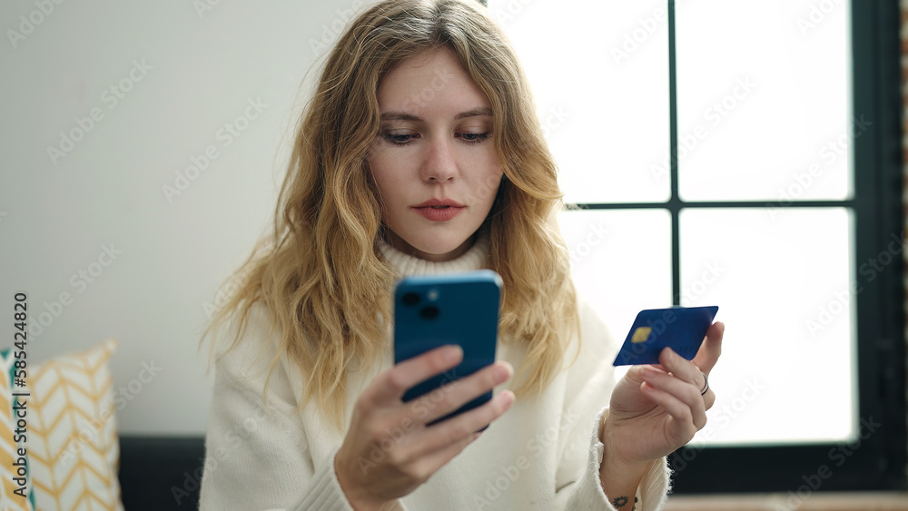 Wall mural young blonde woman using smartphone and credit card sitting on sofa at home