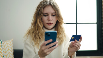 Young blonde woman using smartphone and credit card sitting on sofa at home