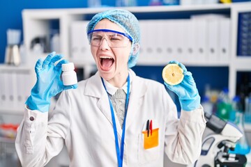 Beautiful woman working at scientist laboratory holding pills and lemon angry and mad screaming frustrated and furious, shouting with anger. rage and aggressive concept.