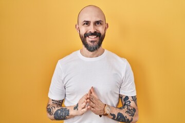 Young hispanic man with tattoos standing over yellow background hands together and fingers crossed smiling relaxed and cheerful. success and optimistic