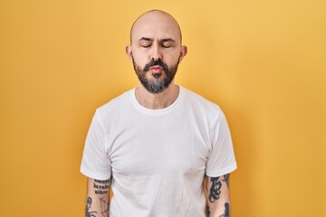 Young hispanic man with tattoos standing over yellow background looking at the camera blowing a kiss on air being lovely and sexy. love expression.