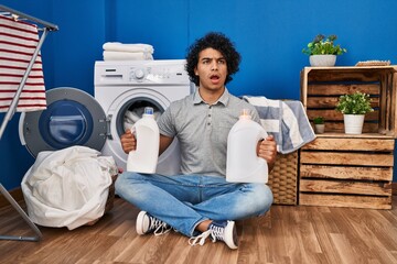 Hispanic man with curly hair doing laundry holding detergent bottles in shock face, looking...