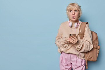 Puzzled worried blonde woman poses with rucksack prepares for hiking trip holds smartphone chats online dressed in casual pullover and pink trousers isolated over blue background copy space.