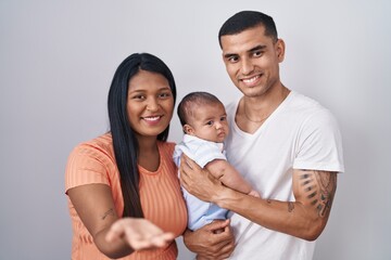 Young hispanic couple with baby standing together over isolated background smiling cheerful offering palm hand giving assistance and acceptance.