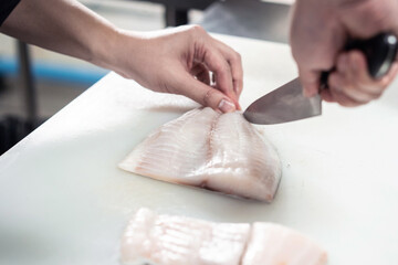 Focused male chef in uniform cutting fish fillet on board, chef cuts white tilapia fish in kitchen.