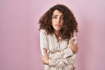 Hispanic woman with curly hair standing over pink background shaking and freezing for winter cold...
