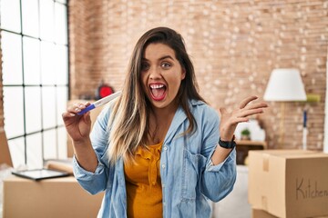 Young hispanic woman holding pregnancy test result at new home celebrating victory with happy smile and winner expression with raised hands