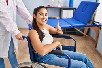Young hispanic woman sitting on wheelchair at physiotherapy clinic surprised with an idea or...