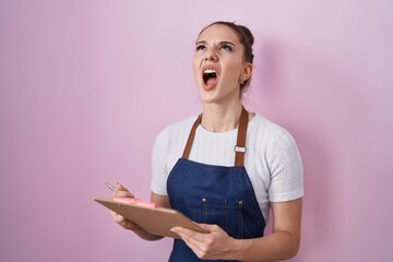 Young hispanic girl wearing professional waitress apron taking order angry and mad screaming...