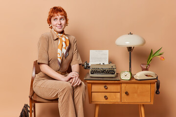 Horizontal shot of redhead retro woman wears elegant costume prepares for job interview looks aside with dreamy expression uses old typewriter and stationary telephone isolated over brown wall