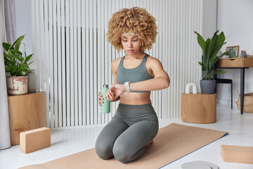Athletic curly haired woman checks time on smartwatch poses at knees on fitness mat dressed in...
