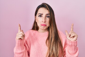 Young hispanic woman standing over pink background pointing up looking sad and upset, indicating...