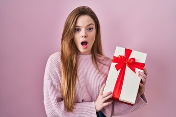 Young caucasian woman holding gift afraid and shocked with surprise and amazed expression, fear and excited face.