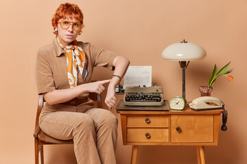 Serious strict redhead European woman indicates at watch asks not to be late dressed in elegant outfit wears big spectacles poses on chair uses retro typewriter isolated over brown background