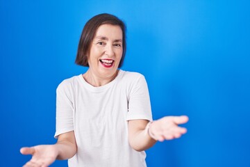 Fototapeta premium Middle age hispanic woman standing over blue background smiling cheerful with open arms as friendly welcome, positive and confident greetings
