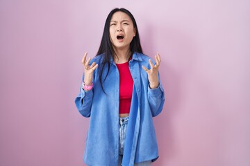 Young asian woman standing over pink background crazy and mad shouting and yelling with aggressive expression and arms raised. frustration concept.