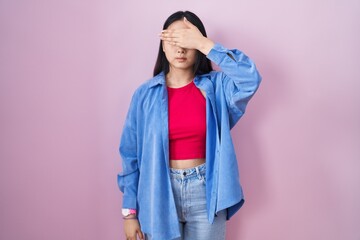 Young asian woman standing over pink background covering eyes with hand, looking serious and sad....