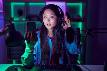 Young asian woman playing video games with smartphone smiling with hand over ear listening an hearing to rumor or gossip. deafness concept.